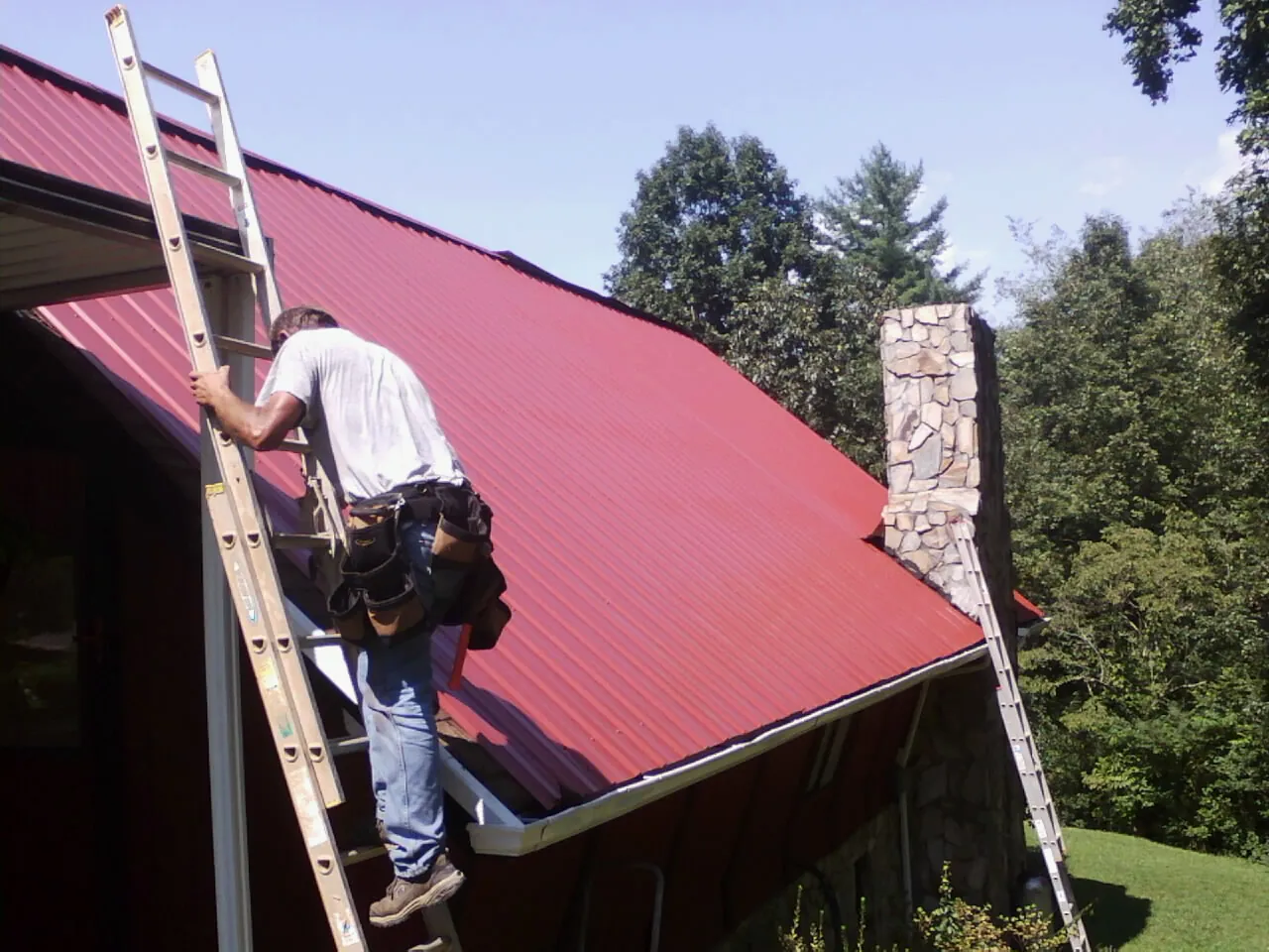 Roofer on a ladder beside a red metal roof and stone chimney — Soffit & Fascia Repair by Roofing Brothers in Milltown
